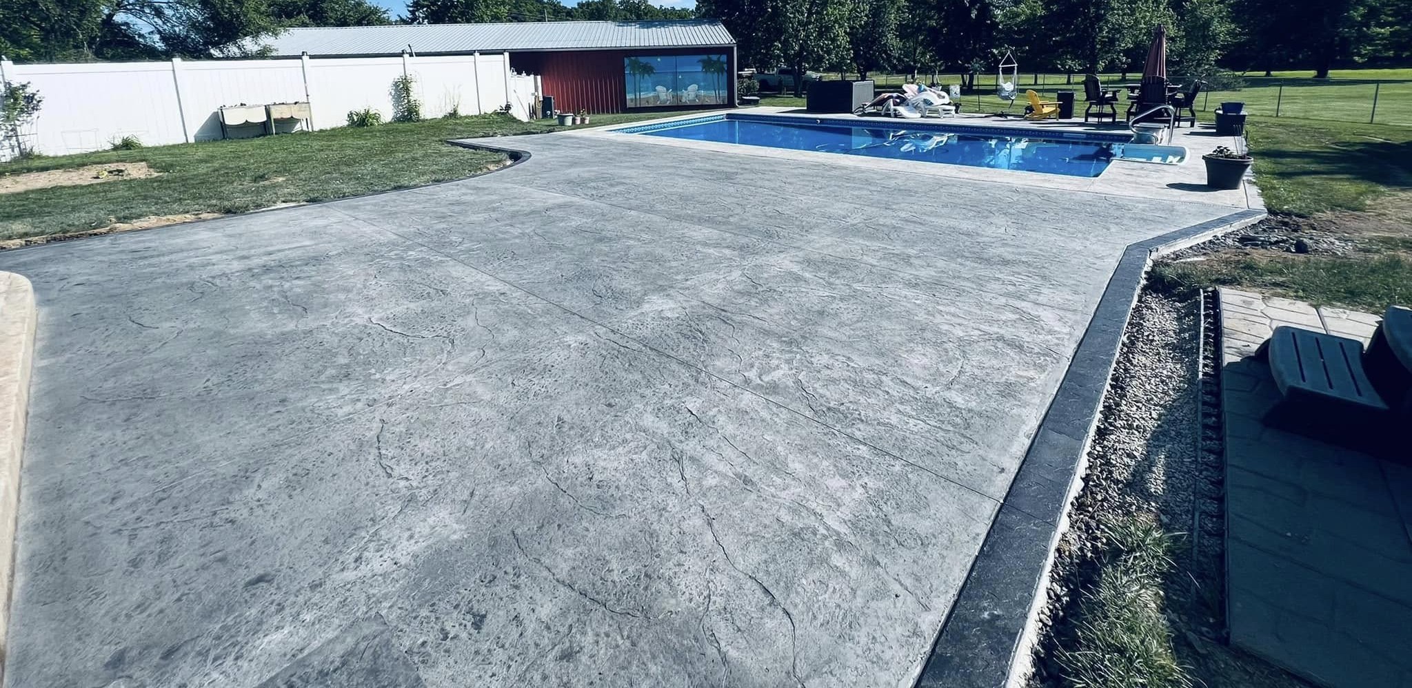 Expansive stamped concrete pool deck in the foreground with an in-ground pool and trees behind